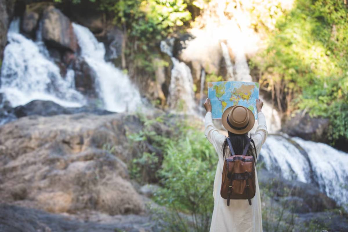 A woman in a hat stands by a waterfall, holding a map up as she explores the scenic surroundings.