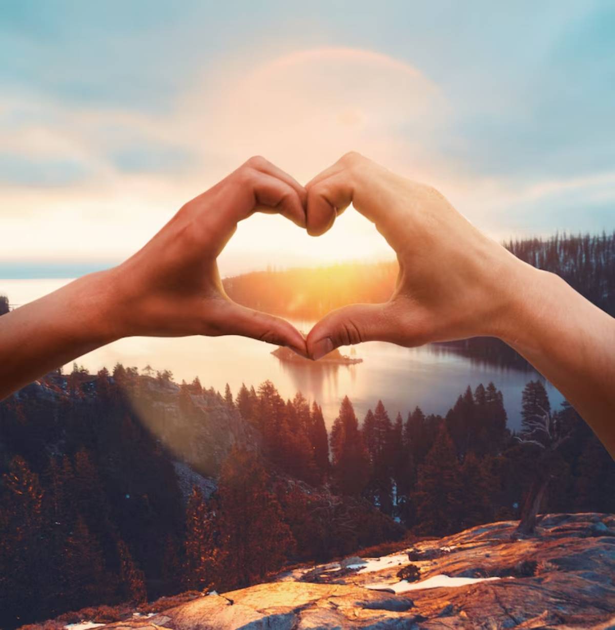 Two hands forming a heart shape against a scenic sunset over a serene lake and forested landscape.