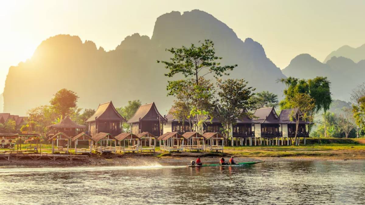 Serene riverside scene featuring traditional wooden houses, lush greenery, and people paddling a boat against a mountain backdrop at sunset.