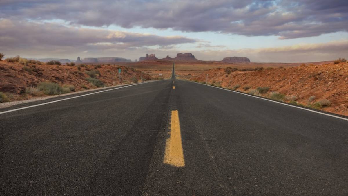 A distant view of a straight road cutting through a desert landscape, leading towards majestic red rock formations under a cloudy sky.