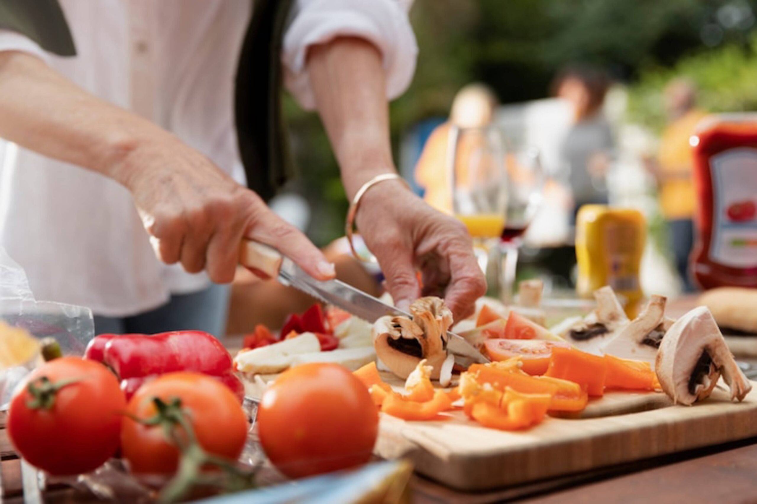 A close-up of a hand slicing vegetables, including tomatoes and peppers, on a wooden cutting board during an outdoor gathering.