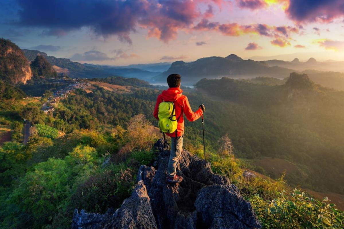 A hiker in a red jacket stands on a rocky outcrop, gazing at a lush green valley under a colorful sunrise sky.