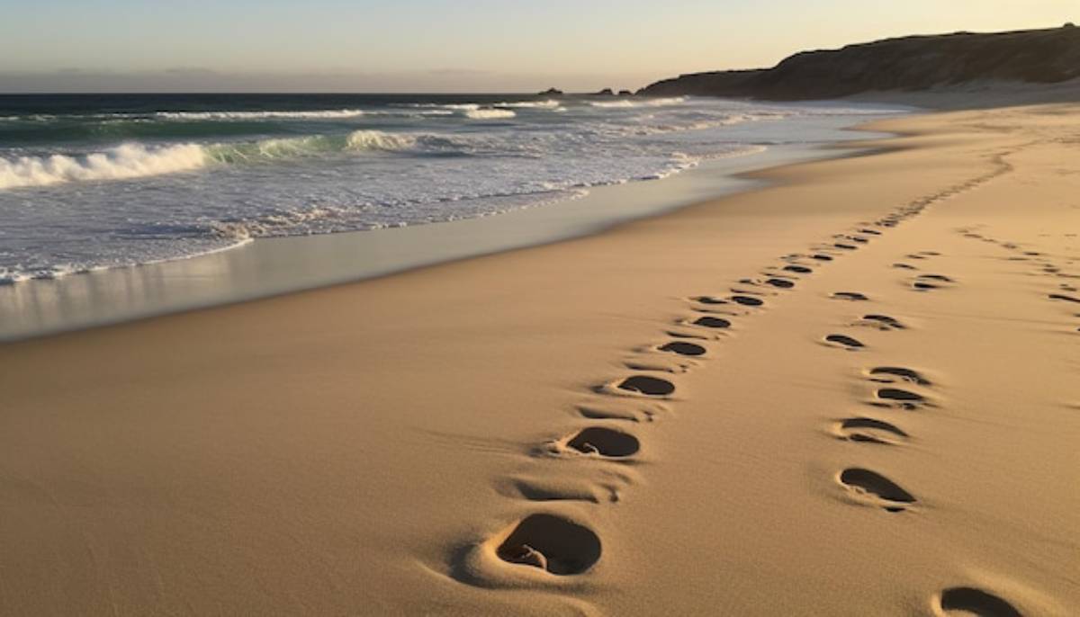 Footprints trail across a sandy beach, leading toward gentle waves under a soft, golden sunset sky.