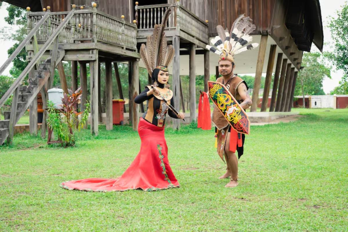 Two individuals in traditional attire perform in front of a large wooden structure, showcasing vibrant costumes and cultural accessories.