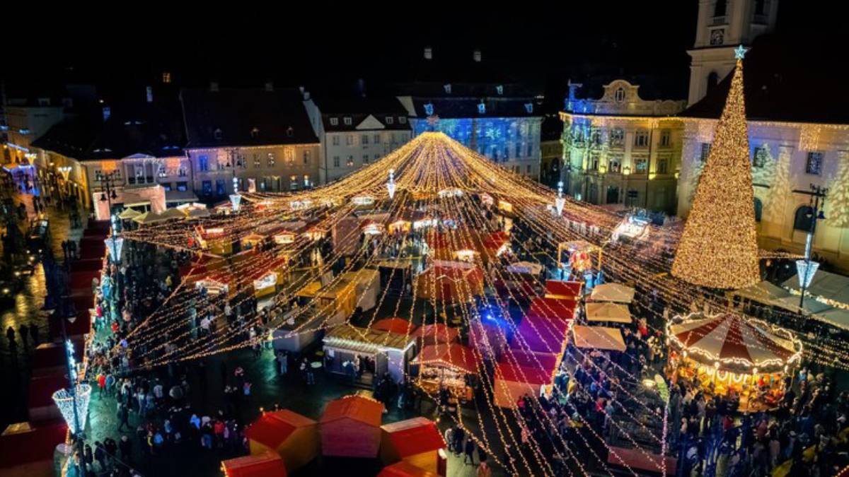 Aerial view of a vibrant Christmas market at night, adorned with lights, stalls, a large decorated tree, and crowds enjoying the festivities.