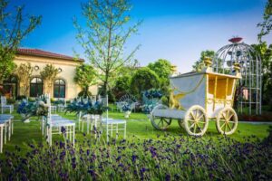 A whimsical outdoor wedding setup featuring a stylish carriage, white chairs, and vibrant floral decorations against a blue sky backdrop.
