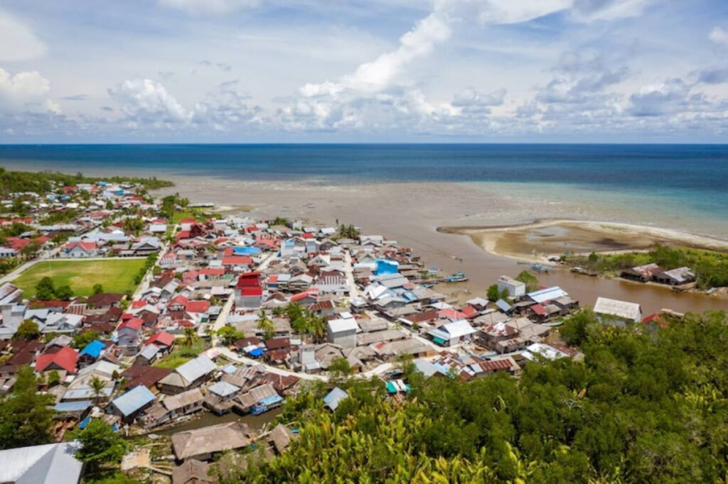 Aerial view of a coastal village with colorful houses, surrounded by lush greenery and a calm sea under a partly cloudy sky.