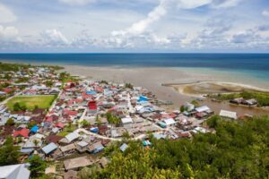 Aerial view of a coastal village with colorful houses, surrounded by lush greenery and a calm sea under a partly cloudy sky.