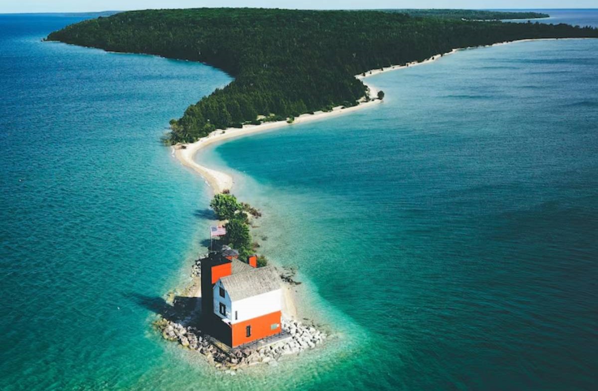 Aerial view of a small red and white lighthouse on a rocky island, surrounded by clear blue waters and a sandy beach at the shore.
