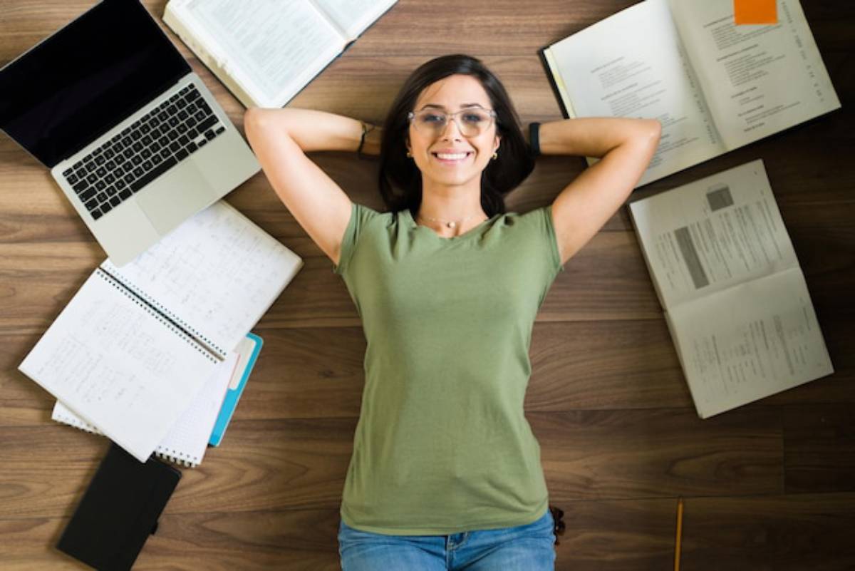 A person lies on the floor surrounded by open books, notebooks, and a laptop, suggesting a study or brainstorming session.