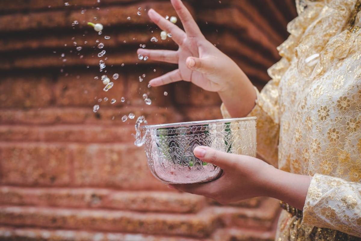 A person in a golden traditional outfit splashes water from a decorative bowl, with droplets sparkling in the air.