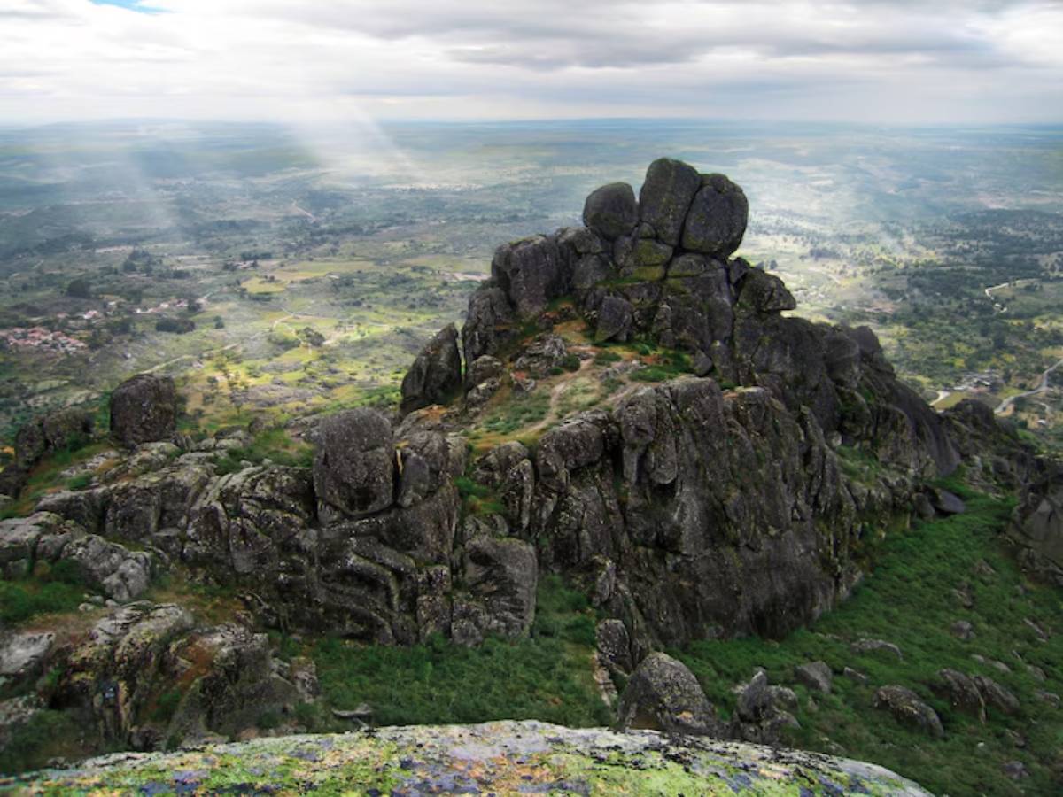A rugged rocky peak rises above lush green valleys under a cloudy sky, with beams of sunlight breaking through the clouds.