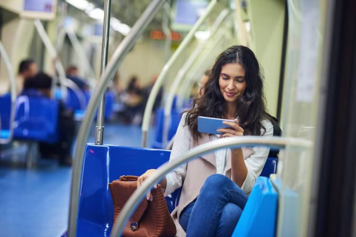 A young woman sits on a blue subway seat, looking at her phone with a brown bag beside her, surrounded by other passengers.