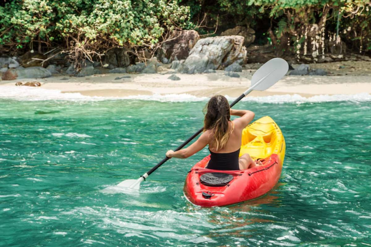 A woman paddles a red kayak in clear turquoise water, heading towards a sandy beach surrounded by lush greenery.