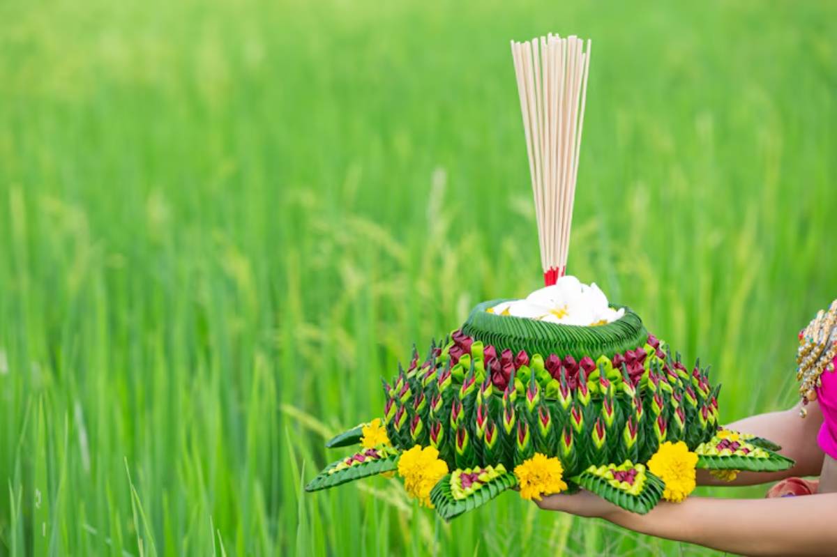 A person holds a beautifully adorned rice offering, decorated with flowers and incense sticks, against a lush green rice field backdrop.