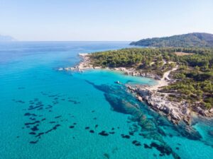 Aerial view of a serene turquoise coastline, bordered by lush greenery and rocky shores under a clear blue sky.