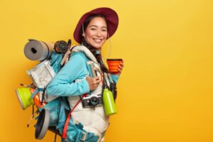 A person in a blue jacket and hat stands against a yellow background, carrying a colorful backpack filled with camping gear and a map.