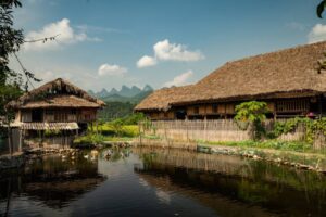 Two traditional thatched-roof houses by a pond, surrounded by lush greenery and mountains under a clear blue sky. Ducks swim in the water.