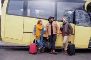 Three travelers stand casually by a yellow bus, holding luggage and drink cups, dressed in stylish, casual winter attire.