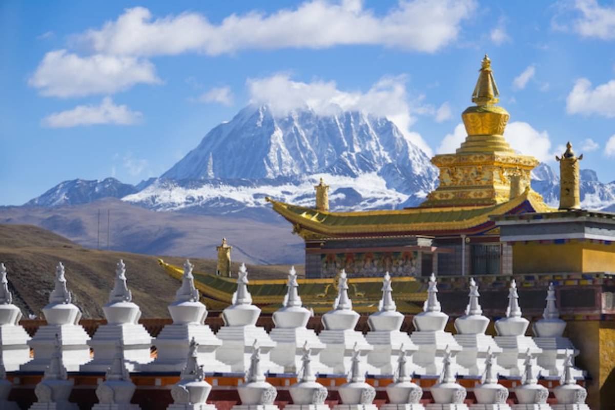 A golden Tibetan temple with intricate architecture stands in front of snow-capped mountains under a bright blue sky.