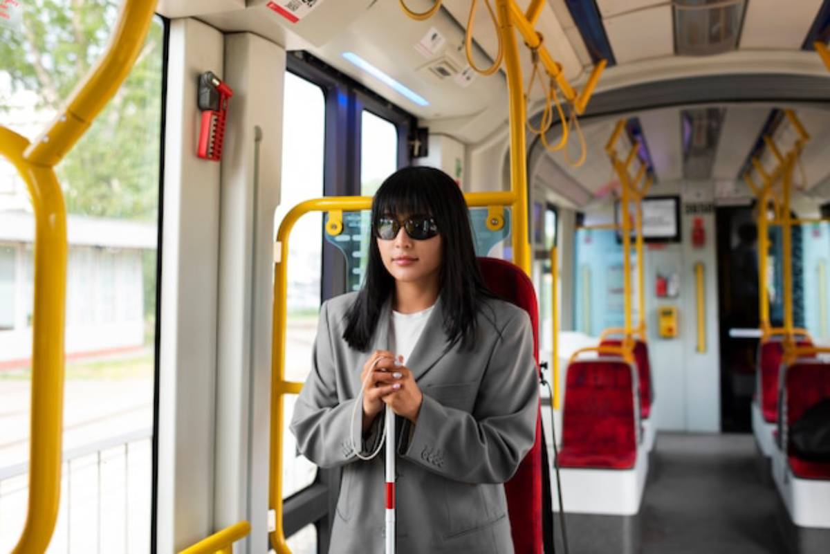 A woman stands inside a bus, holding a white cane, wearing a light gray blazer, surrounded by empty seats and bright yellow handrails.