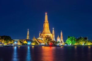 Illuminated Wat Arun temple stands majestically against the night sky, reflected in the water, showcasing intricate architecture.