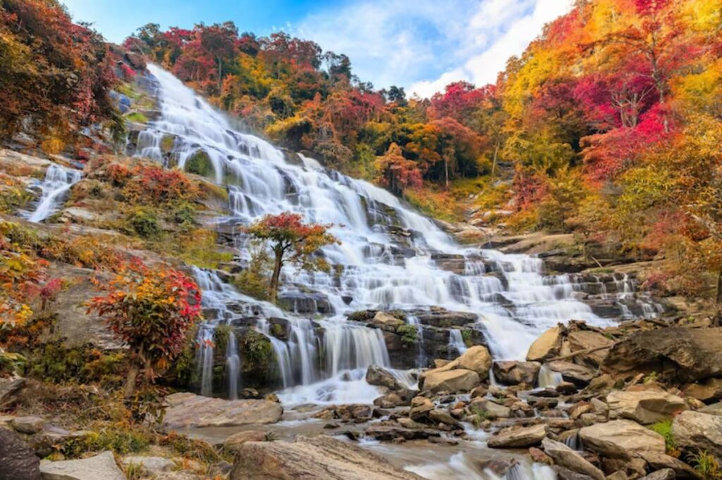 A stunning waterfall cascades down a rocky slope, surrounded by vibrant autumn foliage in shades of red and gold under a partly cloudy sky.