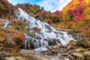 A stunning waterfall cascades down a rocky slope, surrounded by vibrant autumn foliage in shades of red and gold under a partly cloudy sky.