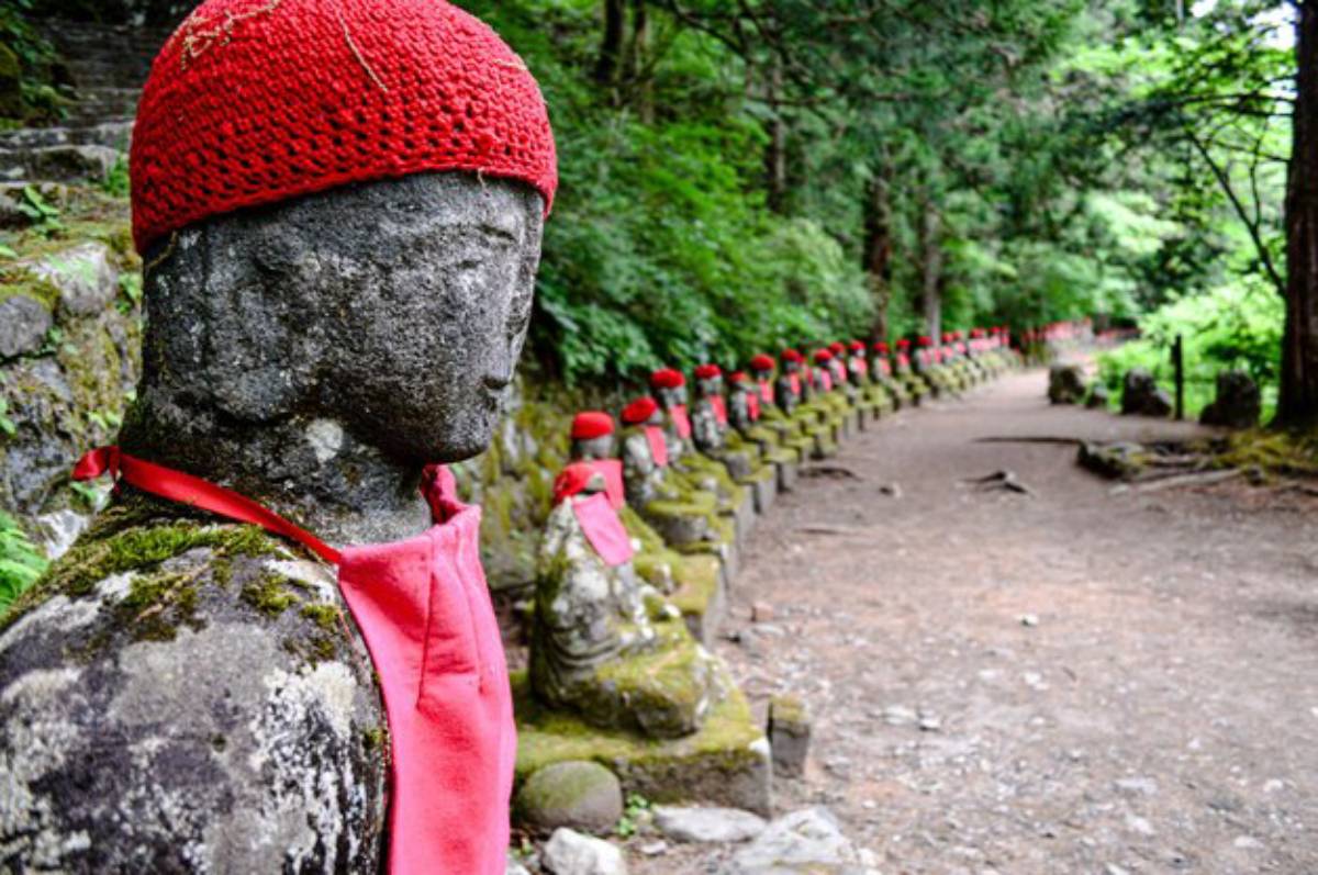 A row of stone statues adorned with red hats and aprons, nestled in a lush green forest setting along a winding path.