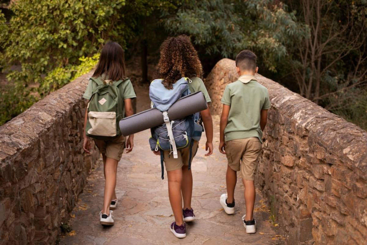 Three children in casual clothing walk together on a stone bridge surrounded by greenery, carrying backpacks and a yoga mat.