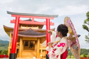 A person in a traditional kimono holds a parasol while pointing at a large red torii gate and a wooden temple in the background.