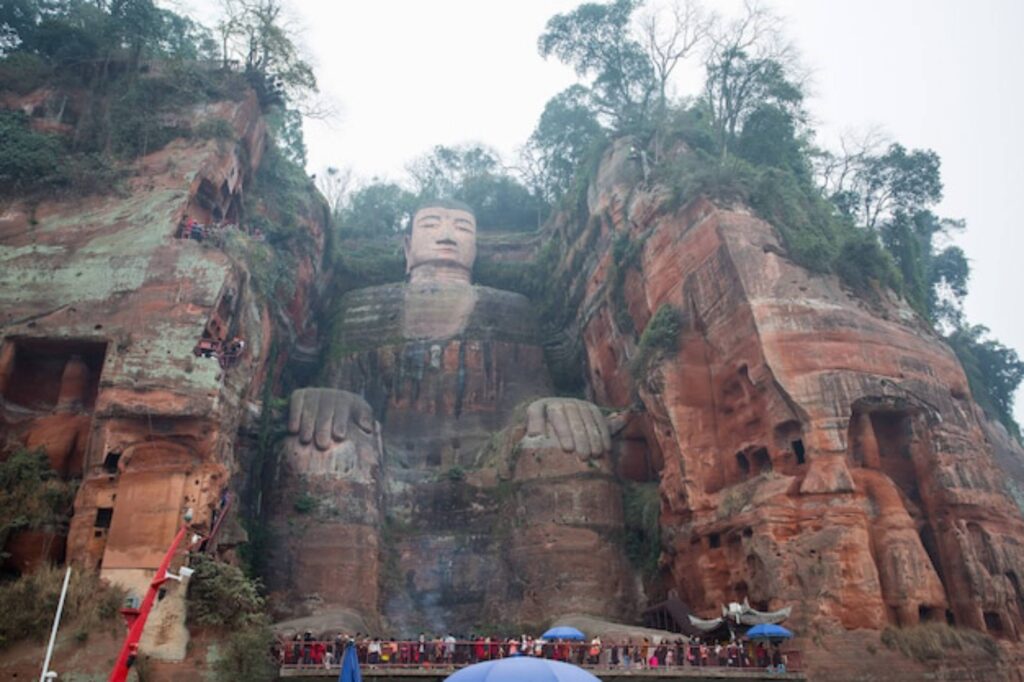 A large stone statue of a seated Buddha with oversized hands, set among rocky cliffs and greenery, with visitors observing below.