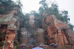 A large stone statue of a seated Buddha with oversized hands, set among rocky cliffs and greenery, with visitors observing below.