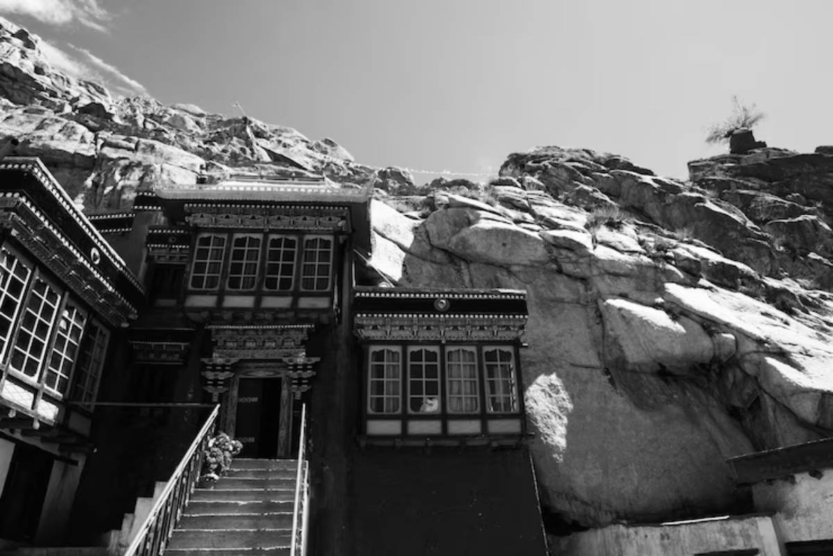 A traditional Tibetan building nestled against rugged rock formations, captured in black and white. Stairs lead to its ornate entrance.