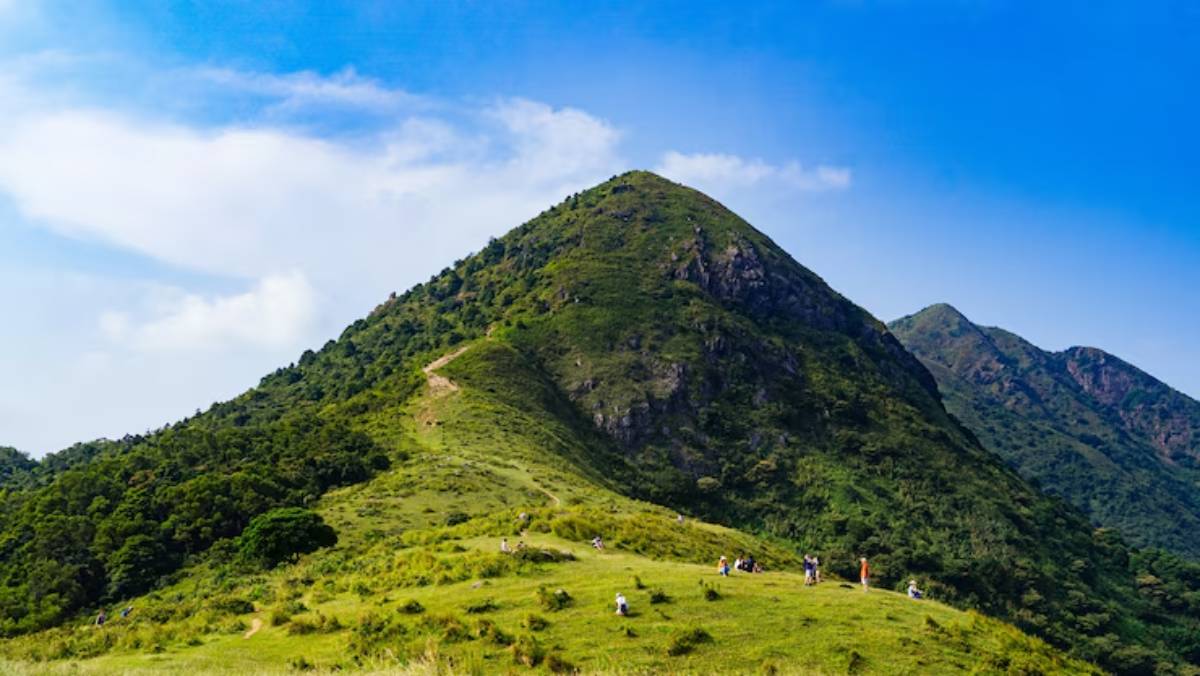 A picturesque mountain landscape with hikers on grassy slopes, under a bright blue sky with wispy clouds.
