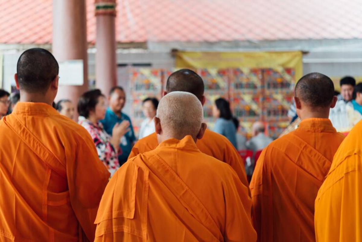 A group of monks in vibrant orange robes stands with their backs to the camera, observing a lively gathering in a temple setting.