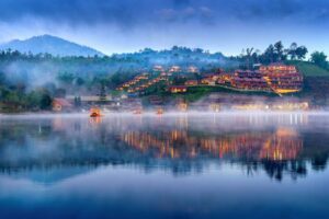 A serene lakeside village at dawn, with mist rising and colorful lights reflecting on the water, surrounded by lush mountains.