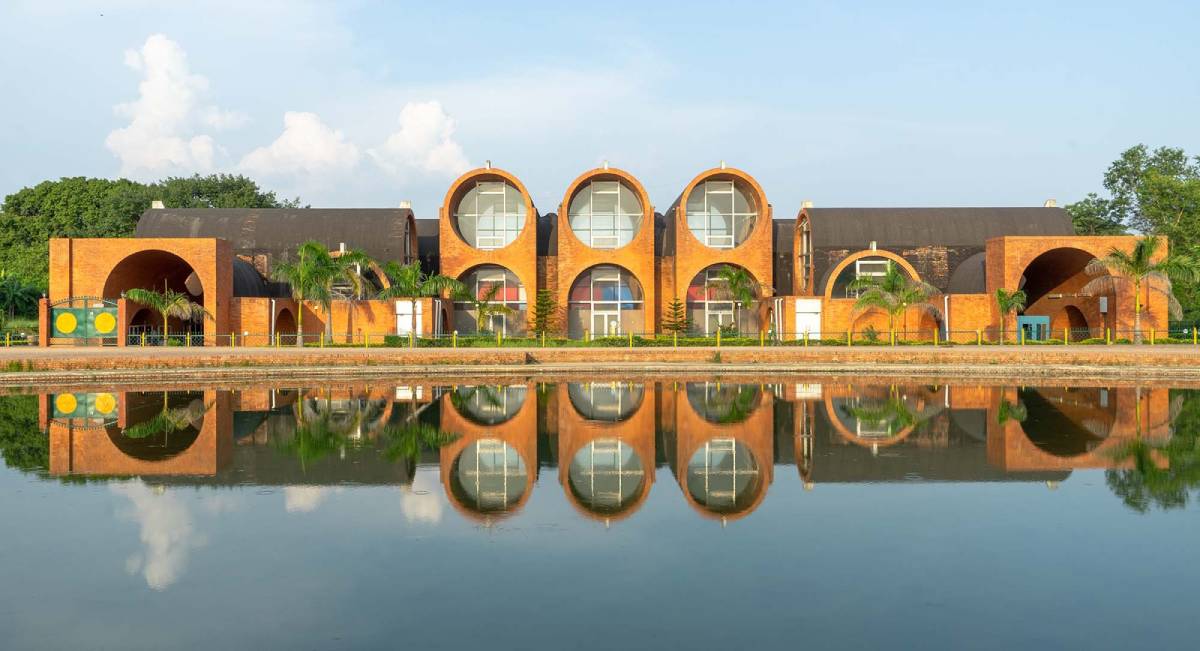 Modern brick building with large circular windows, reflecting in a serene pond under a blue sky with scattered clouds.