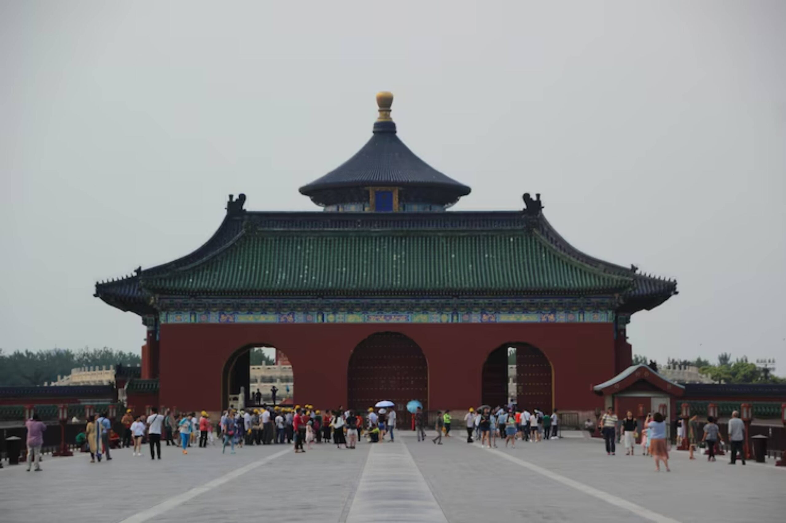 A crowd gathers at the iconic Temple of Heaven in Beijing, featuring its distinct green and blue roof under an overcast sky.