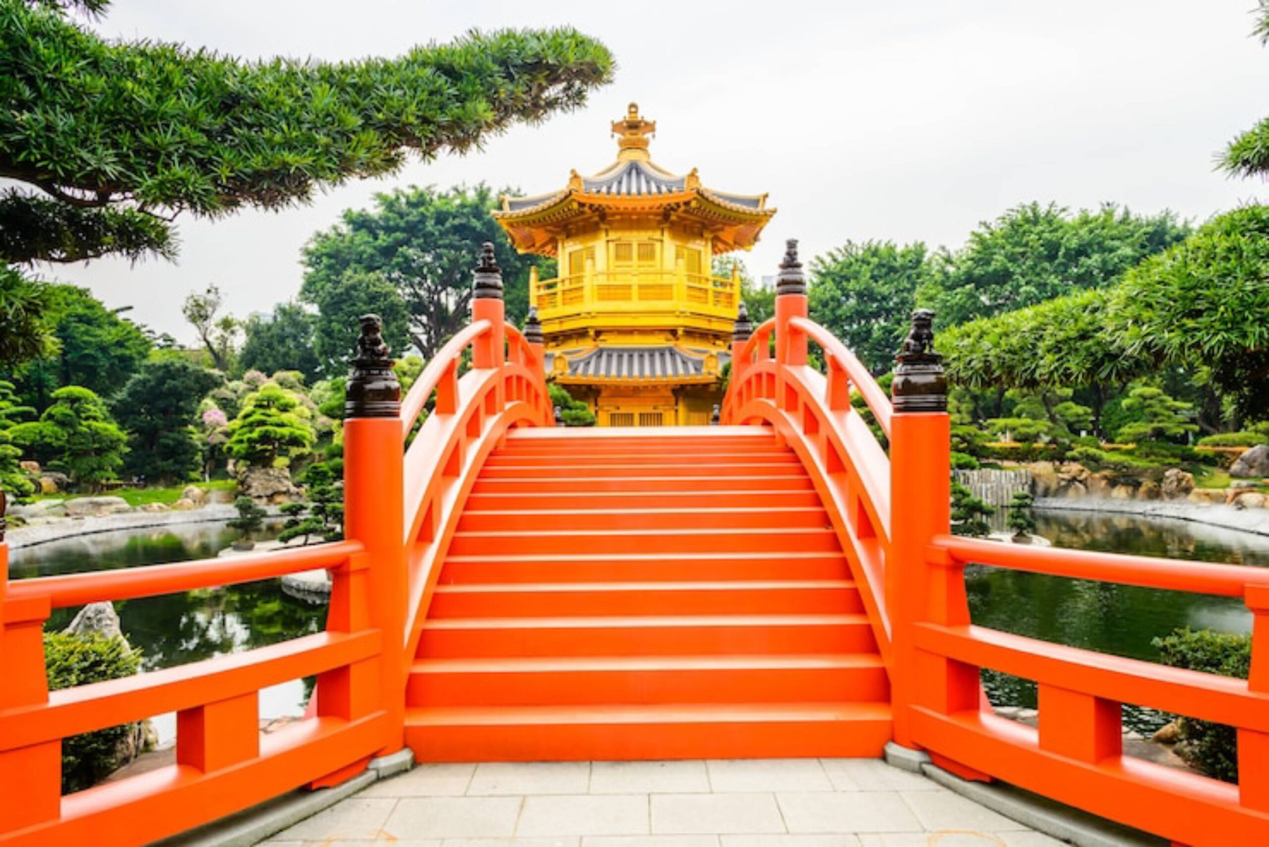 A vibrant orange bridge leads to a golden pavilion surrounded by lush greenery and a serene pond in a tranquil garden setting.