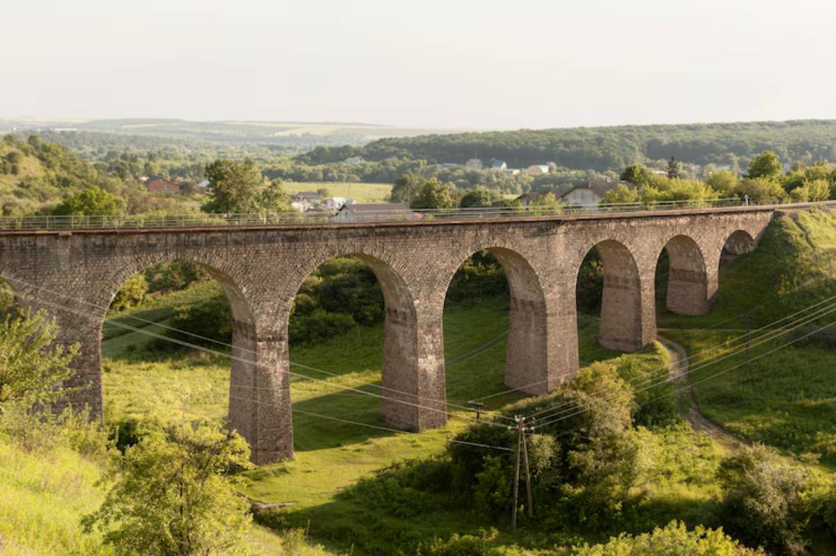 A historic stone viaduct with multiple arches spans lush green hills under a clear sky, with scattered homes in the background.