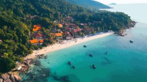 Aerial view of a tropical beach resort with orange-roofed buildings, sandy shore, and boats on clear turquoise water surrounded by lush greenery.