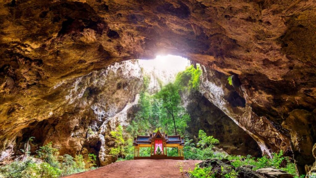 A vibrant temple sits beneath a rocky cave, surrounded by lush greenery and illuminated by sunlight filtering through the opening above.