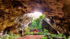 A vibrant temple sits beneath a rocky cave, surrounded by lush greenery and illuminated by sunlight filtering through the opening above.
