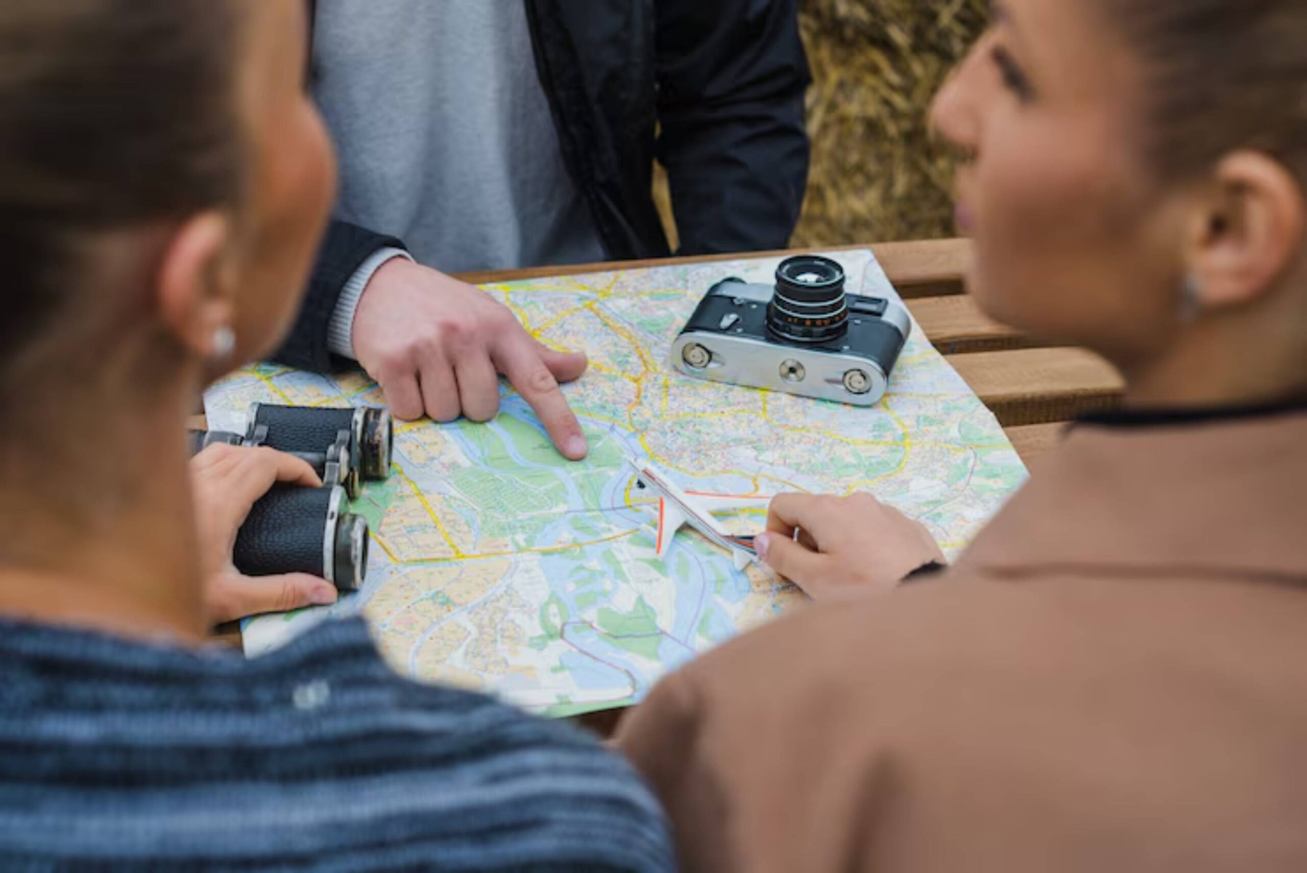 Two people examining a detailed map on a table, with binoculars and a vintage camera nearby, indicating planning or exploration.