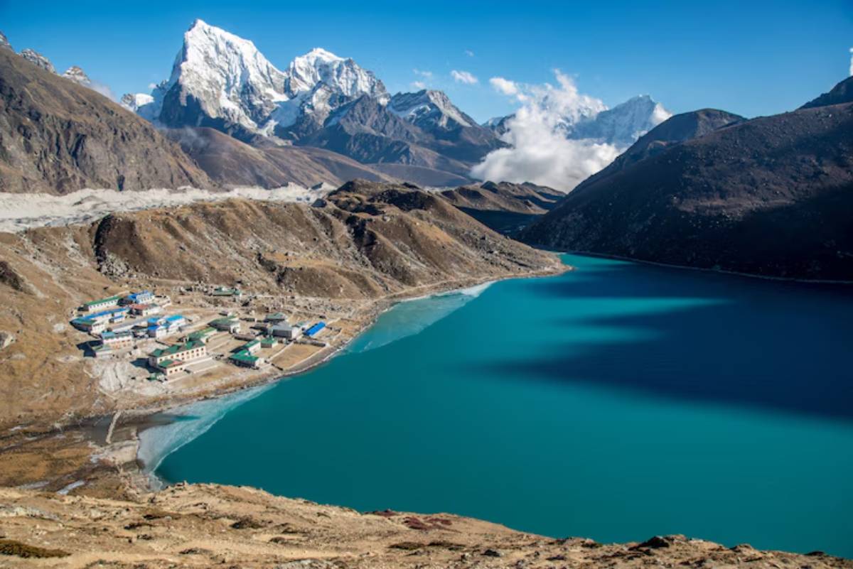 A scenic view of a turquoise lake surrounded by rugged mountains and a small village with colorful buildings under a clear blue sky.