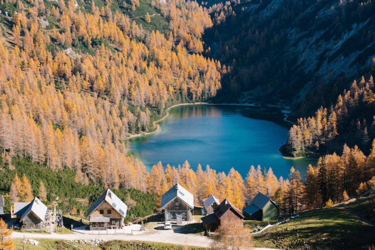 Scenic view of quaint wooden cabins beside a serene blue lake, surrounded by autumn-colored larch trees in the mountains.