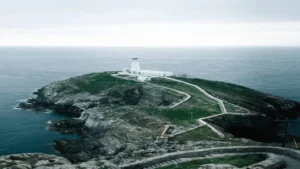 A lighthouse stands atop a rugged cliff, surrounded by the sea, with a winding path leading to it under a cloudy sky.