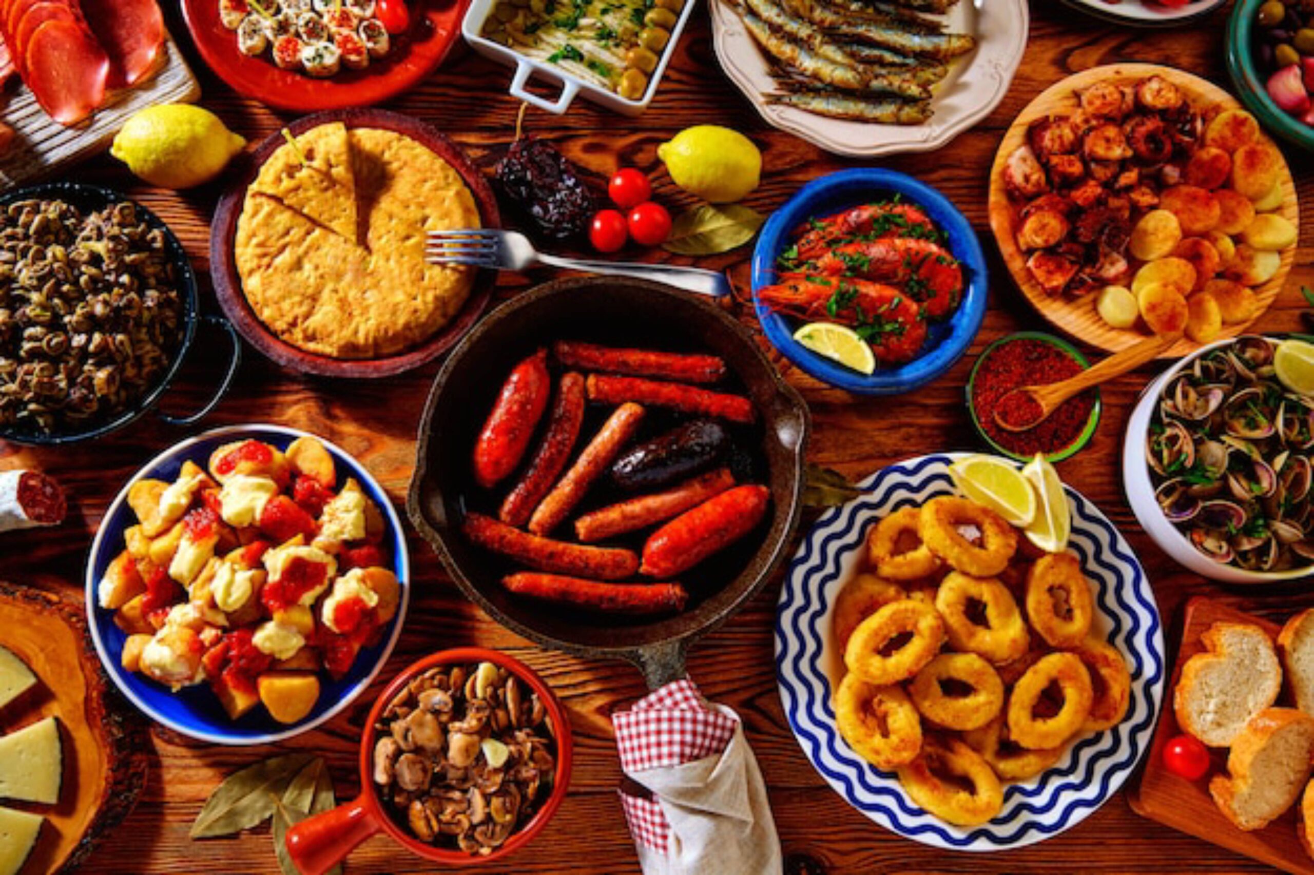 A vibrant spread of assorted dishes: grilled sausages, fried calamari, seafood, potatoes, cheese, and various appetizers on a rustic table.