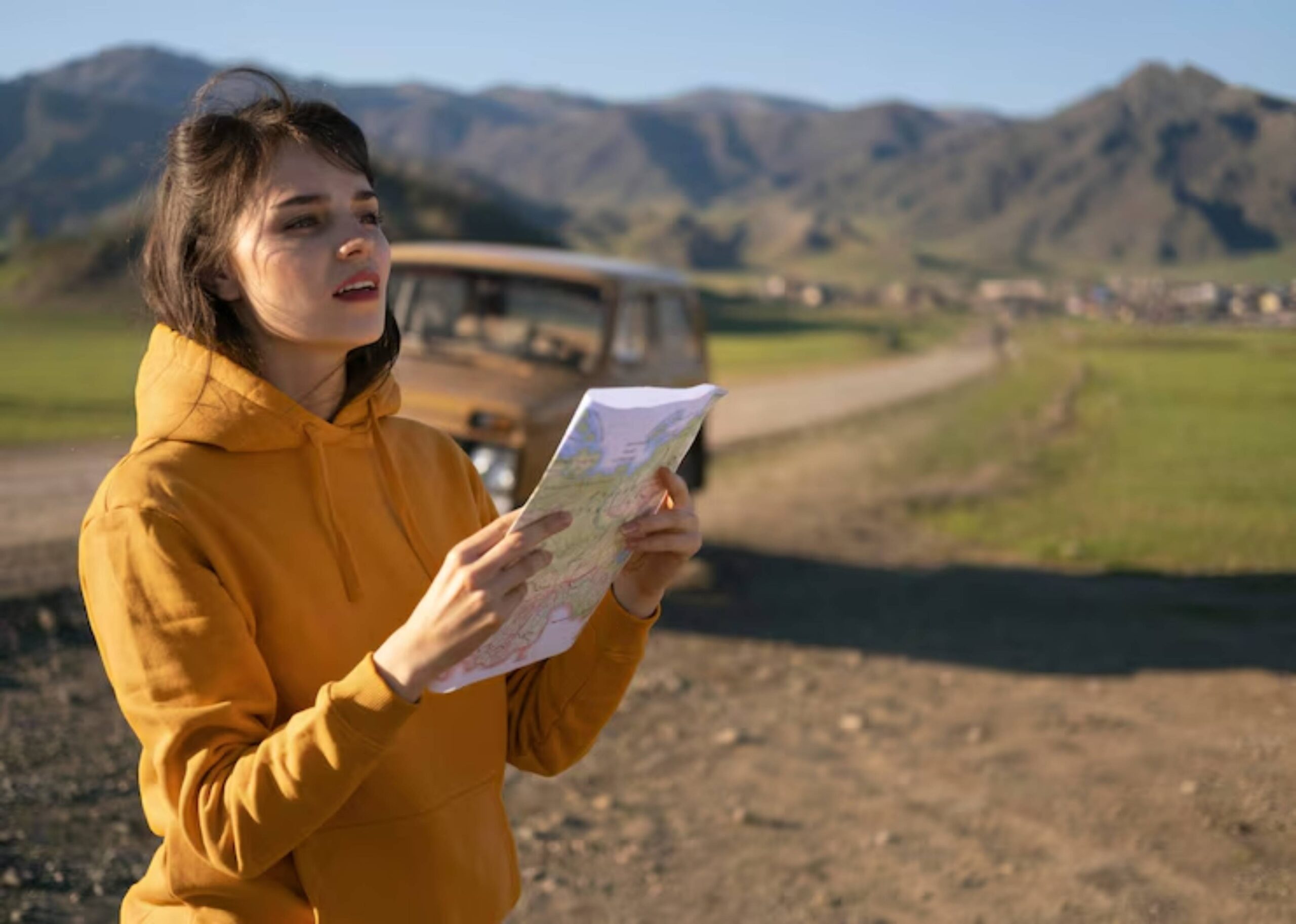 A person in a bright orange hoodie studies a map in a scenic, mountainous landscape, with an old vehicle parked nearby.